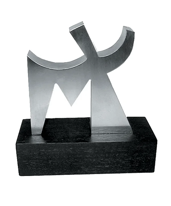 A 6” polished stainless steel abstract “M” sculpture mounted on a black wooden stand